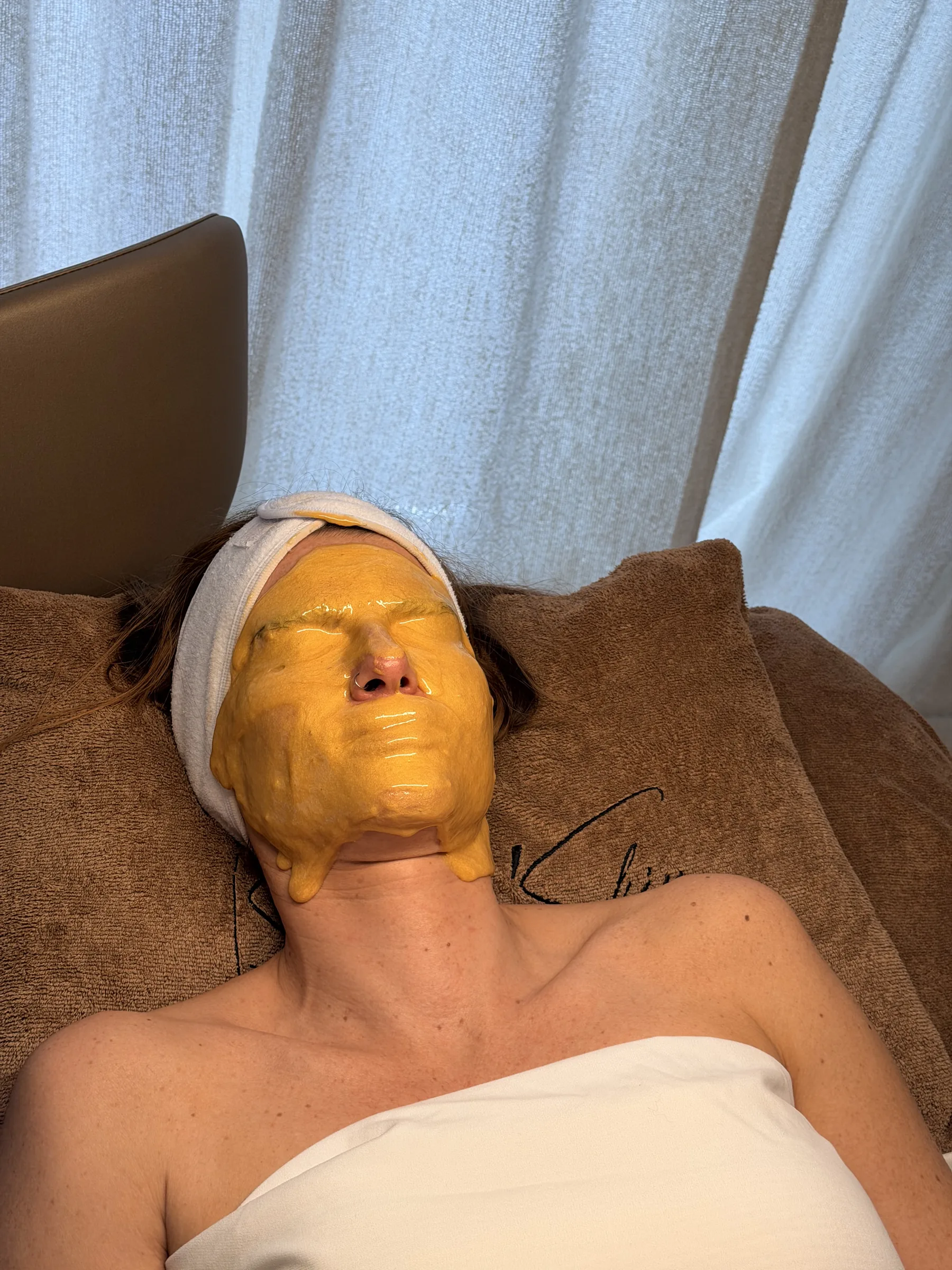 Klant ligt met een oranje gelmasker en een witte haarband op de behandeltafel