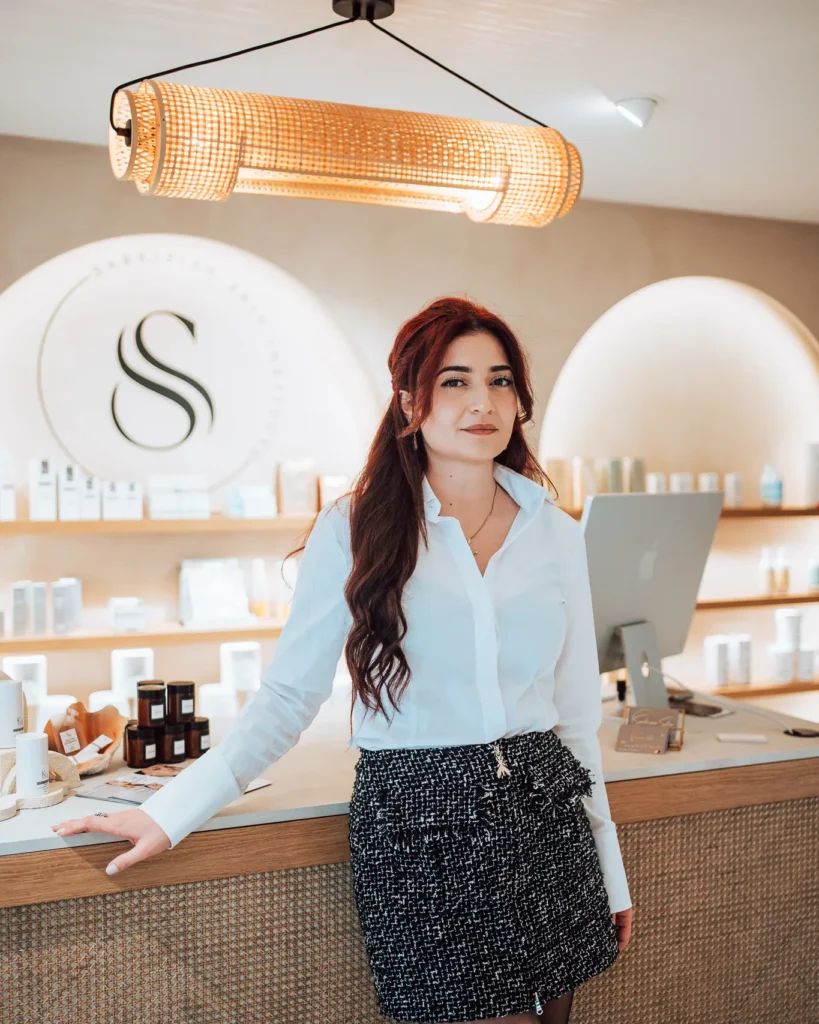 Marin Sarkisian leunt tegen de onthaalbalie van haar salon, met het verlichte S-logo en de productnissen achter haar