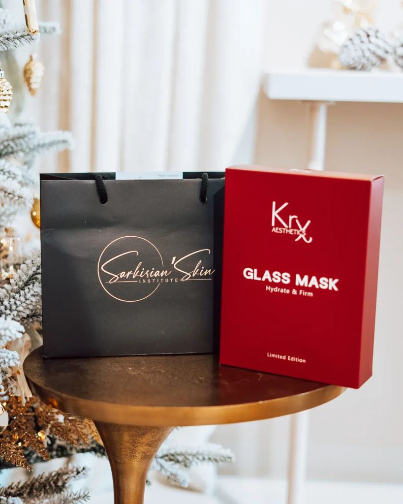 Zwarte geschenktas van Sarkisian Skin Institute naast een doos KRX Glass Mask op een gouden bijzettafel bij de kerstboom